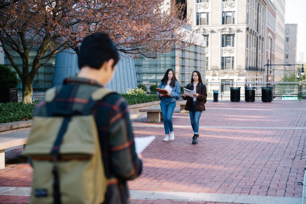 College students carrying notebooks and backpacks walking across a paved campus courtyard in the city